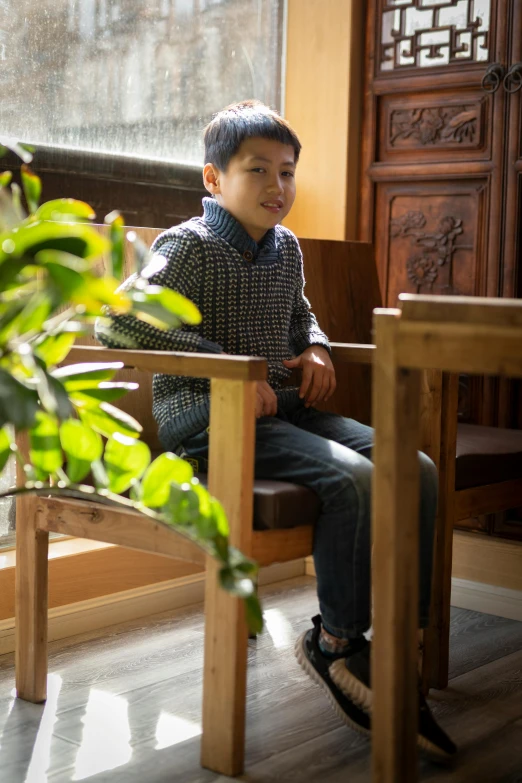 a young boy sitting on a bench next to a potted plant, sitting on a mocha-colored table, kakar cheung, profile image, seated on wooden chair