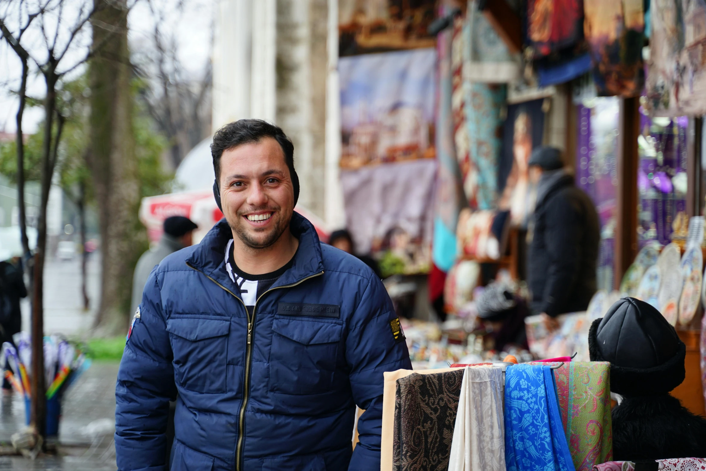 a man in a blue jacket standing in front of a market, an album cover, pexels contest winner, hurufiyya, fabrics and textiles, smiling at the camera, professional profile picture, georgic