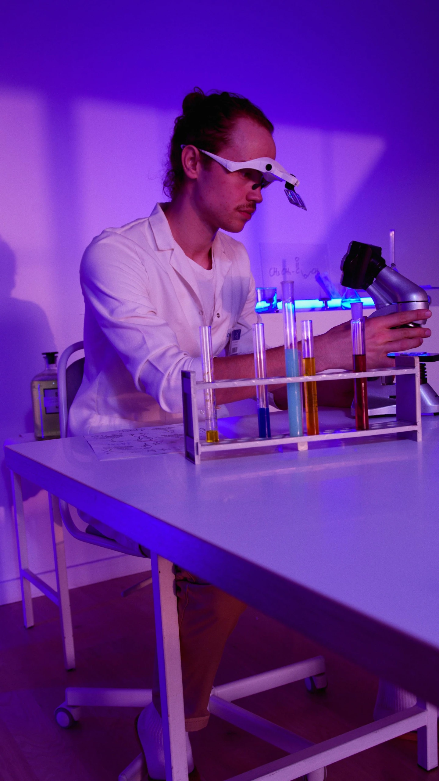 a man sitting at a table in front of a microscope, purple volumetric lighting, chemical woekshop, performance, low quality photo