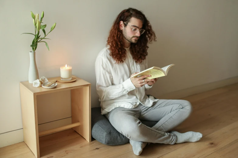 a man sitting on the floor reading a book, by Alice Mason, light and space, wooden side table, meditating pose, on a candle holder, grey