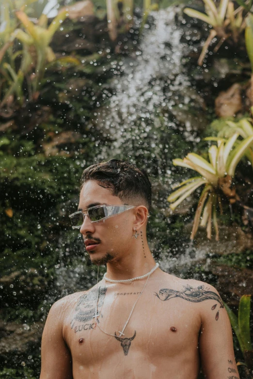 a man standing in front of a waterfall, an album cover, inspired by Randy Vargas, trending on pexels, non binary model, wearing shades, wearing shipibo tattoos, standing in a botanical garden