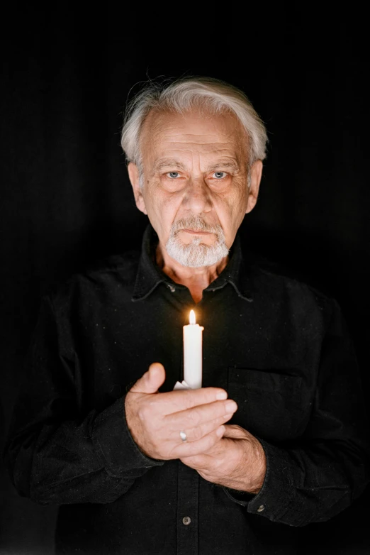 a man holding a lit candle in his hands, an album cover, inspired by Lajos Vajda, an oldman, looking towards the camera, with a beard and a black shirt, solemn gesture