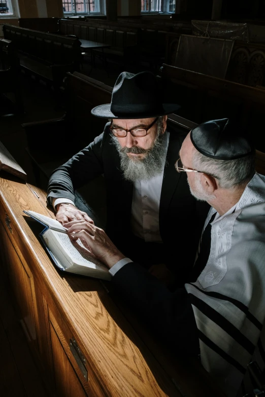 a couple of men sitting next to each other in a room, by Elias Goldberg, pexels, holy ceremony, wizard reading a directory, jewish young man with glasses, a suited man in a hat