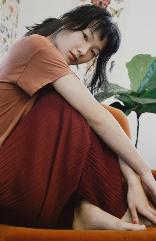 a woman sitting on top of a couch next to a plant, by Tan Ting-pho, trending on pexels, pleated skirt, wearing an orange t-shirt, korean women's fashion model, dull red