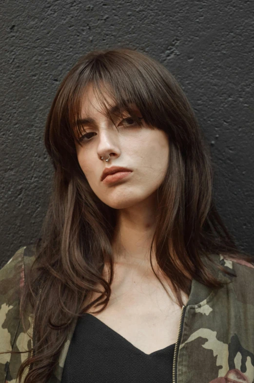 a woman standing in front of a black wall, inspired by Elsa Bleda, trending on unsplash, photorealism, brown long hair with bangs, non binary model, militaristic, ana de armas portrait