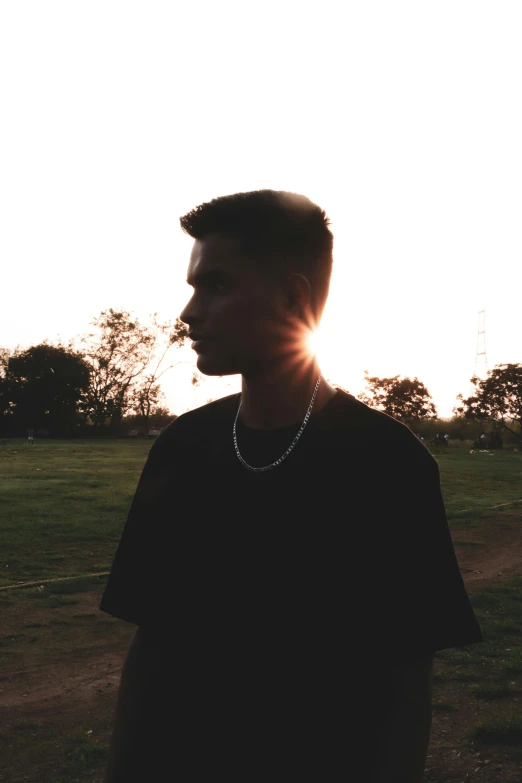 a man in a black shirt standing in a field, an album cover, unsplash, realism, ((sunset)), slightly defined jawline, at a park, slightly tanned