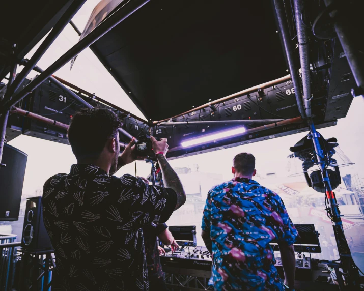 a couple of men standing next to each other in front of a stage, unsplash, process art, dj set, sitting in a crane, the back rooms, looking down on the camera