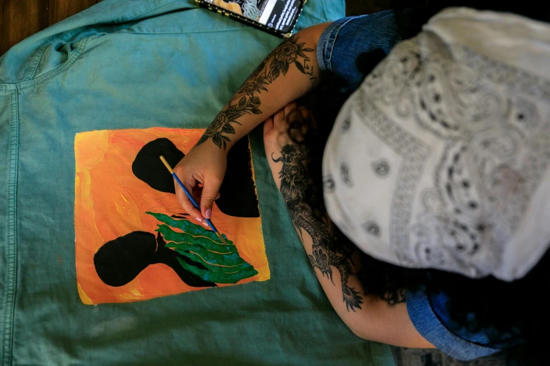 a person sitting on a bed writing on a piece of paper, a painting, featured on pexels, process art, aboriginal australian hipster, teal and orange colours, costume design made with love, stencil