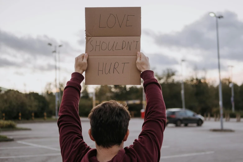 a man holding up a sign in a parking lot, by Julia Pishtar, trending on pexels, hurufiyya, love hate love, injured, a broad shouldered, hongbsws