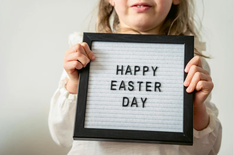 a little girl holding a sign that says happy easter day, trending on unsplash, 1 6 x 1 6, light box, grey, teacher