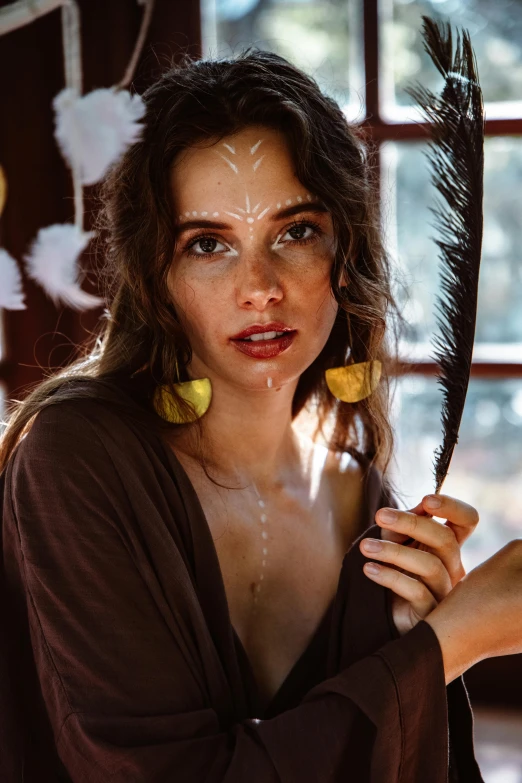 a woman holding a feather in front of a window, a portrait, trending on pexels, forehead jewelry, brunette fairy woman, ocher details, tribal paint
