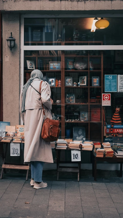 a woman standing in front of a book store, by Matija Jama, pexels contest winner, light brown trenchcoat, modest, facing away, crafts and souvenirs