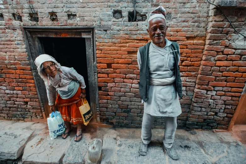 a man and a woman standing in front of a brick building, inspired by Steve McCurry, pexels contest winner, hyperrealism, nepal, bags on ground, portrait photo of an old man, background image