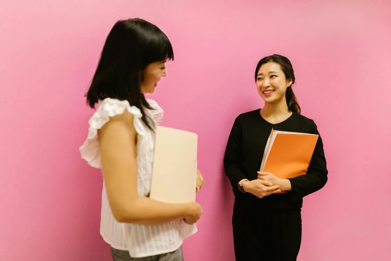 two women standing next to each other in front of a pink wall, by Ayako Rokkaku, academic art, holding notebook, coworkers, low quality photo, reaching out to each other