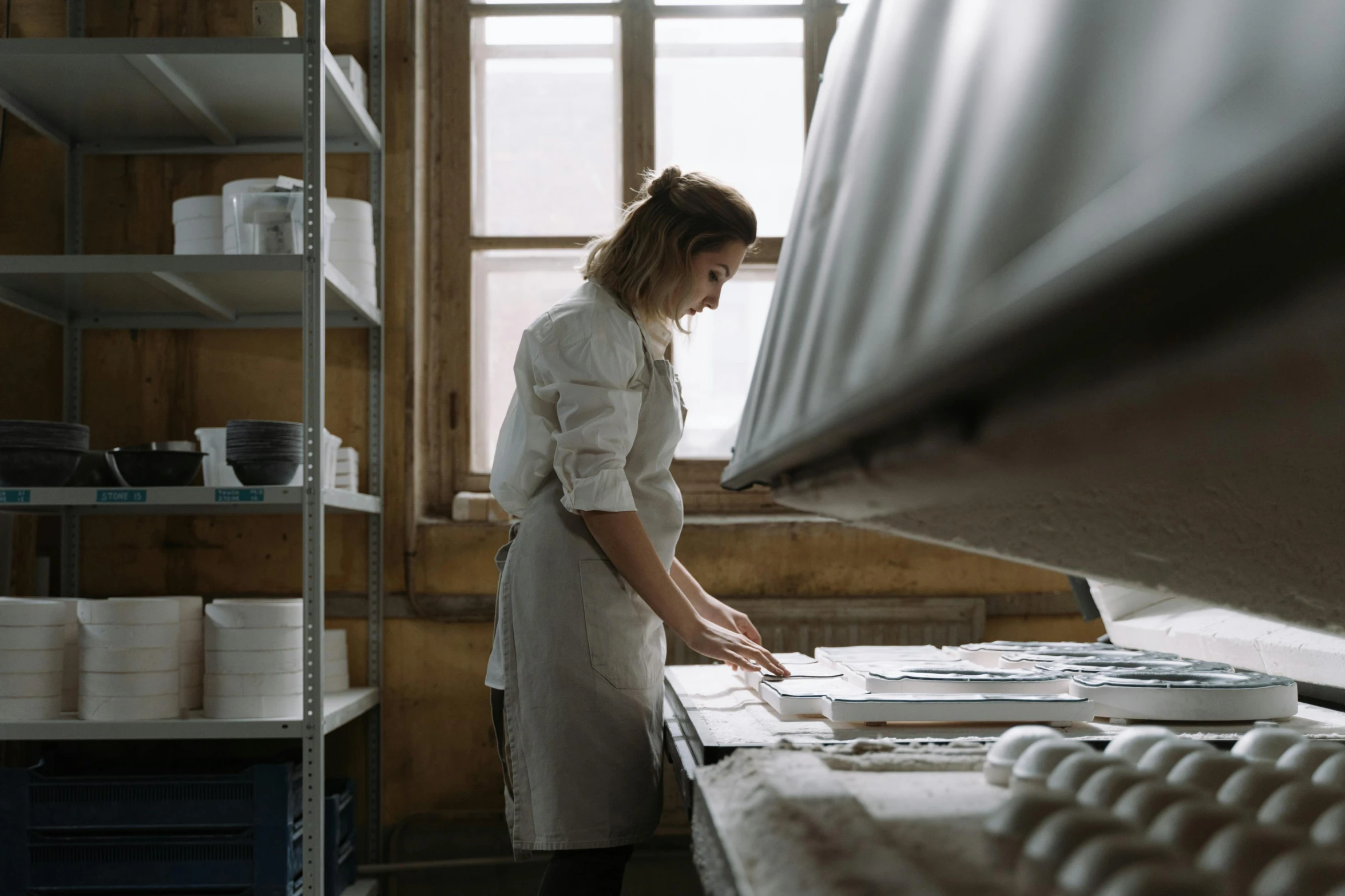 a woman standing in a kitchen preparing food, a silk screen, pexels contest winner, arbeitsrat für kunst, in a warehouse, white ceramic shapes, bakery, looking to the side