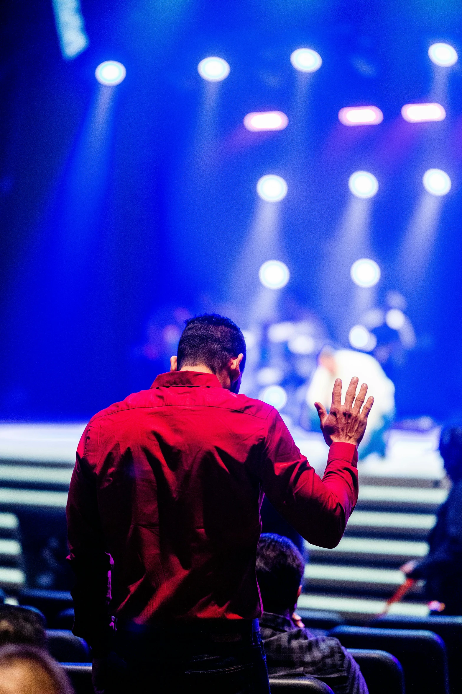 a man in a red shirt standing in front of a stage, a picture, by Dan Content, pexels, happening, kneeling in prayer, holy spirit, facing away from the camera, colours