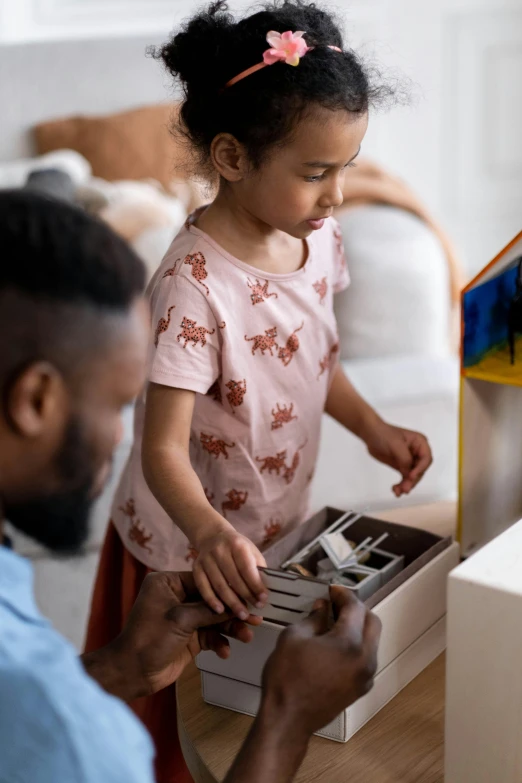a little girl standing next to a man in a living room, interactive art, looking at the treasure box, diverse ages, thumbnail, 2019 trending photo