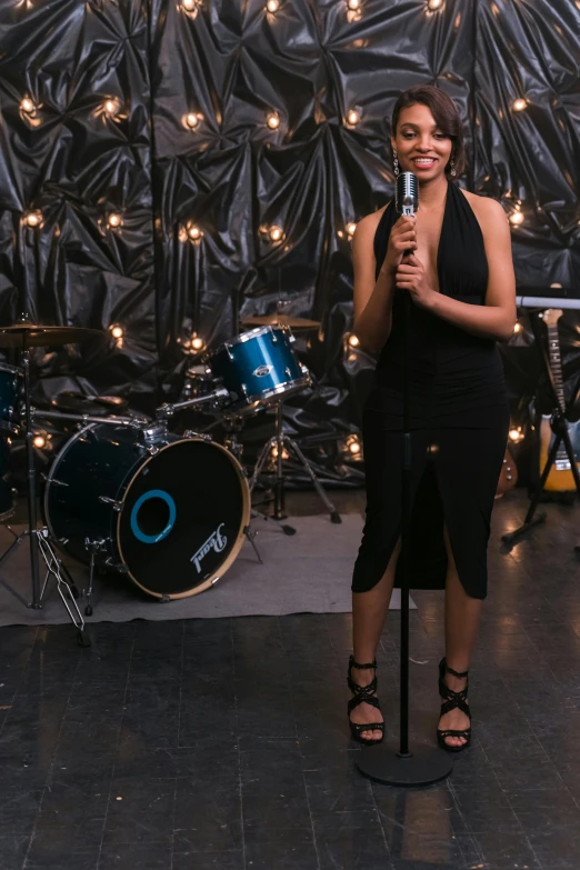 a woman in a black dress holding a microphone, drum set, tessa thompson inspired, panoramic shot, tall shot