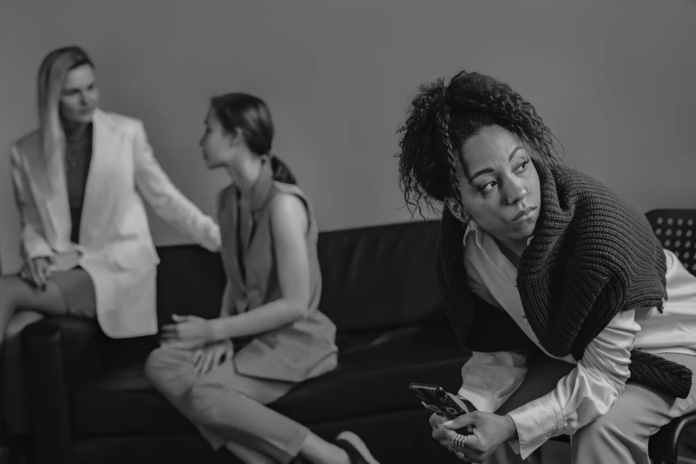 a black and white photo of two women sitting on a couch, by Adam Marczyński, pexels, bullying, african american woman, in an office, people walking around