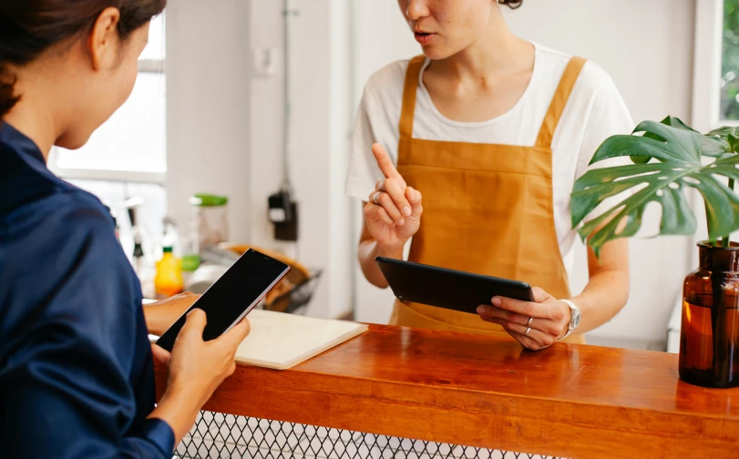 a woman standing in front of a counter holding a tablet, by Lee Loughridge, trending on pexels, medium shot of two characters, serving suggestion, a wooden, bo chen