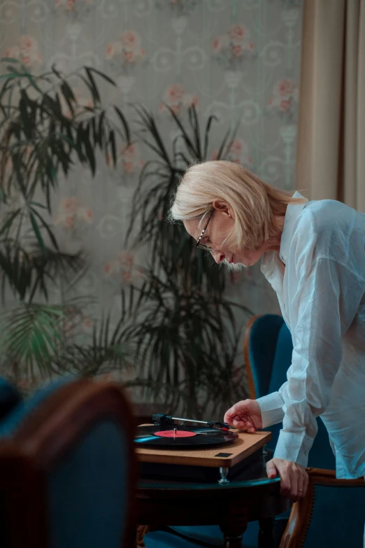 a woman leaning over a table in a living room, an album cover, inspired by Elsa Bleda, trending on unsplash, process art, white-haired, djing with dj turntables, dementia, cardistry