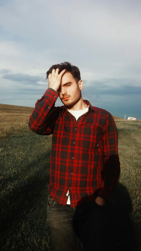a man standing on top of a grass covered field, an album cover, by Robbie Trevino, pexels contest winner, realism, wearing a plaid shirt, light stubble with red shirt, seaside, asher duran