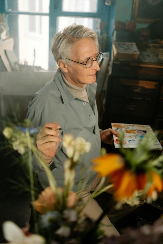 a man sitting at a table with flowers in front of him, pexels contest winner, visual art, holding a paintbrush, wearing medium - sized glasses, frank gehry, avatar image