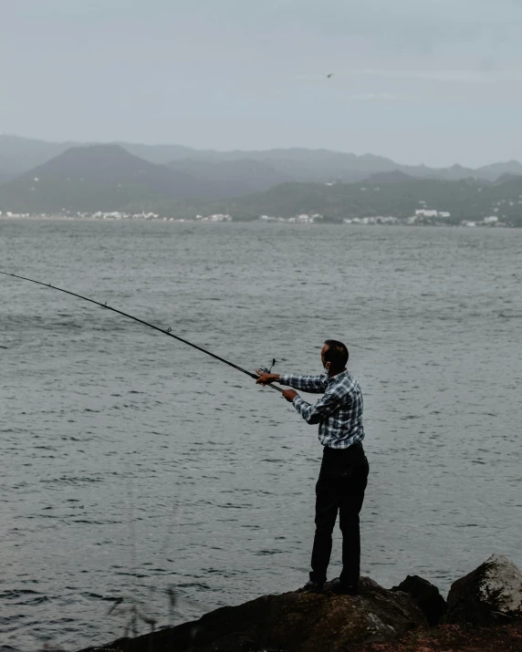a man standing on top of a rock next to a body of water, fishing pole, grey skies, caught on camera, mullet