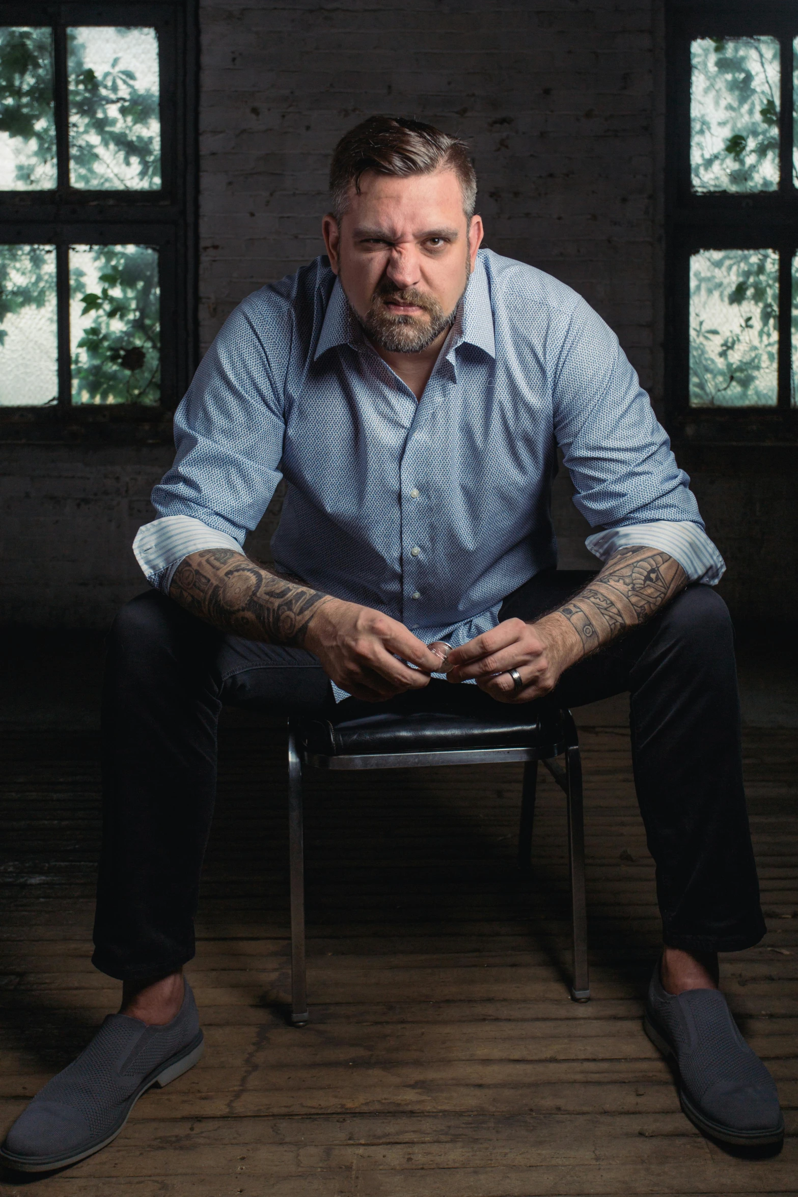 a man sitting on a chair in front of a window, a portrait, by Brian 'Chippy' Dugan, with tattoos, paul giamatti, colour photo, promotional image