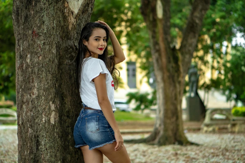 a woman leaning against a tree in a park, by Luis Miranda, pexels contest winner, tachisme, wearing denim short shorts, avatar image, brazilan supermodel, cute young woman