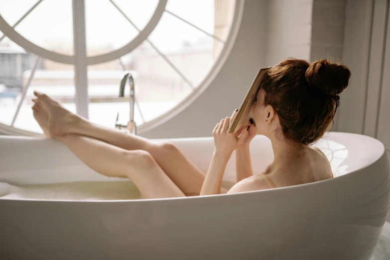 a woman sitting in a bath tub next to a window, trending on pexels, very thin, smooth golden skin, trying to read, sitting in a lounge