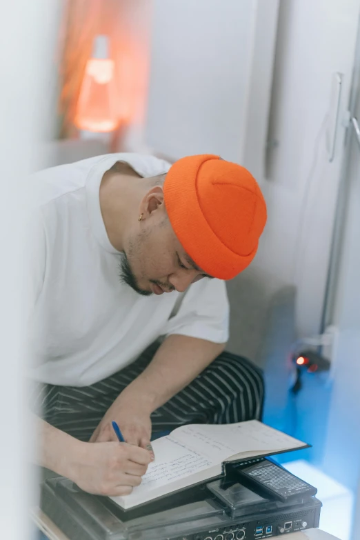 a man in an orange hat writing on a piece of paper, in white room, japanese streetwear, lgbtq, praying