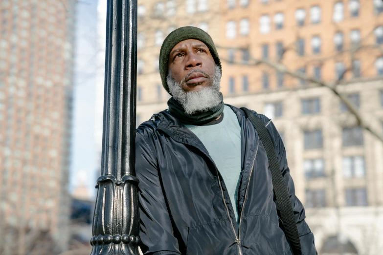 a man standing next to a lamp post with a building in the background, inspired by Gordon Parks, he also wears a grey beanie, humans of new york, [ theatrical ], concerned expression