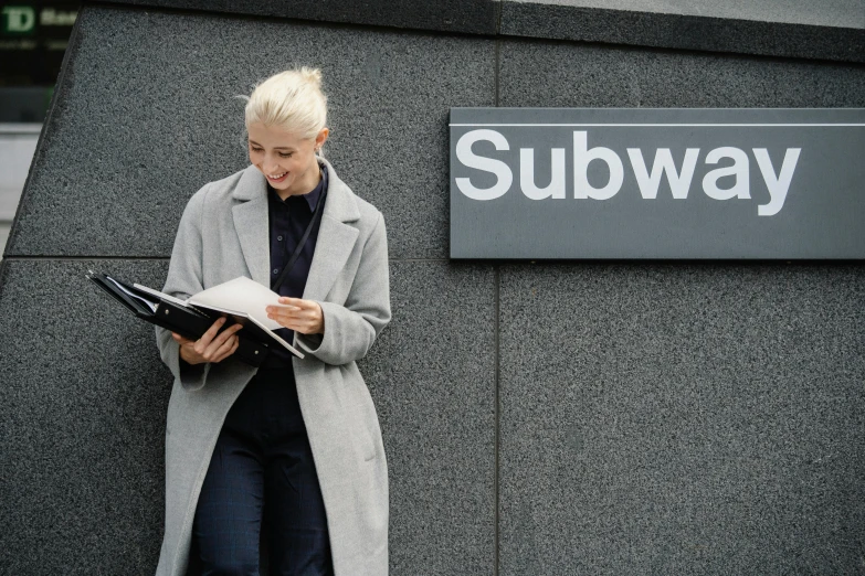 a woman standing in front of a subway sign, unsplash, holding notebook, architect, suburban, a blond