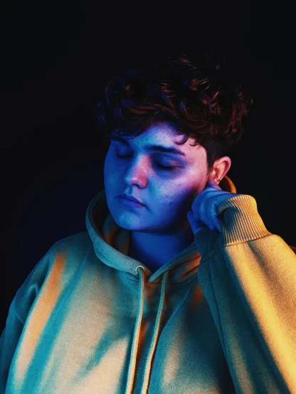 a close up of a person wearing a hoodie, an album cover, inspired by Elsa Bleda, trending on pexels, realism, andy milonakis, god. dramatic gold blue lighting, non binary model, thoughtful pose