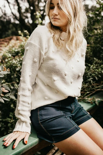 a woman sitting on top of a green bench, wearing a white sweater, with soft bushes, wearing a cute top, round-cropped