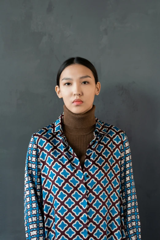 a woman standing in front of a gray wall, inspired by Kim Tschang Yeul, renaissance, blue turtleneck, patterned clothing, brown shirt, demna gvasalia