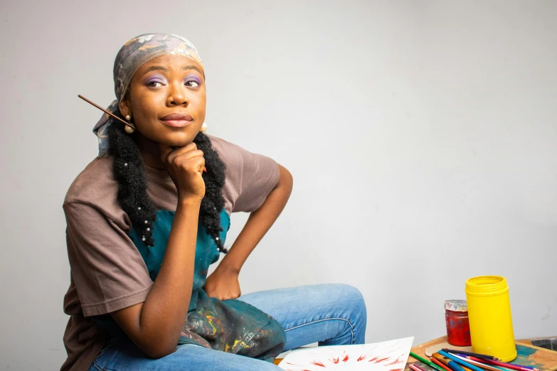 a woman sitting on top of a table holding a paintbrush, by Chinwe Chukwuogo-Roy, pexels contest winner, thinking pose, student, childish look, riyahd cassiem