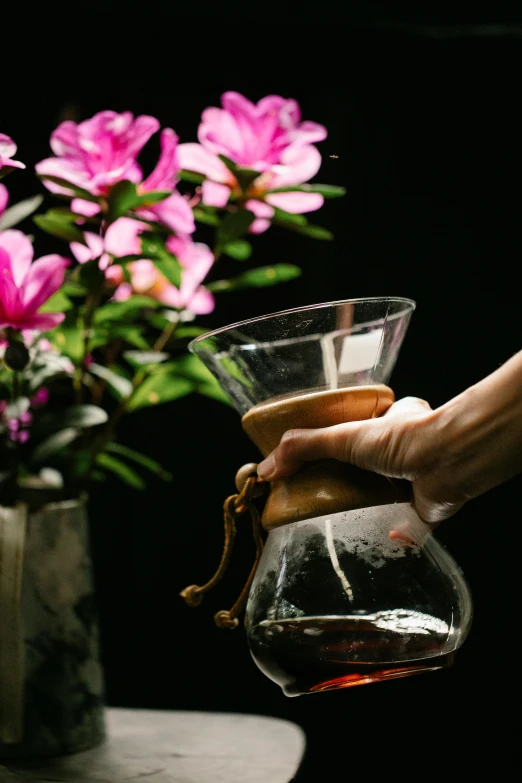 a person holding a coffee pot in front of a vase of flowers, inspired by Elsa Bleda, unsplash, on black background, a wooden, pouring, medium close up