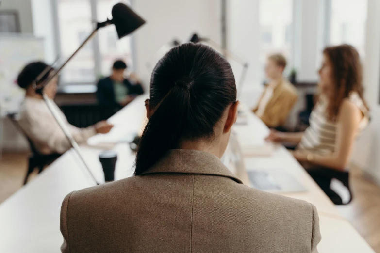 a woman sitting at a table in front of a group of people, trending on pexels, back of head, office clothes, viewed from a distance, thumbnail