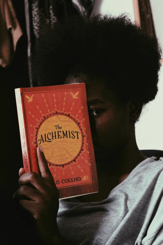 a person holding a book in front of their face, tachisme, african steampunk alchemist, black teenage girl, instagram post, archillect