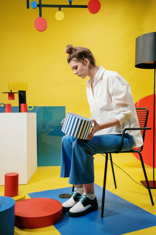 a woman sitting on a chair in a brightly colored room, an album cover, inspired by Bauhaus, trending on unsplash, de stijl, stripey pants, panoramic anamorphic, the woman holds more toys, box