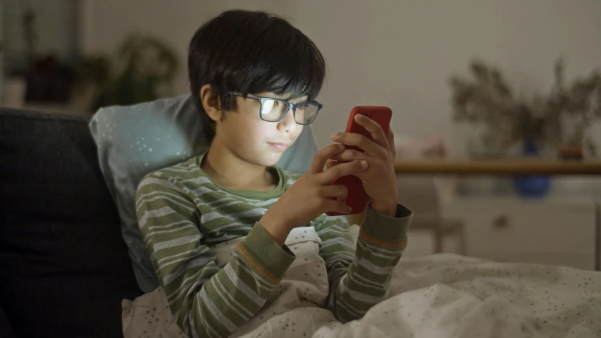 a young boy sitting on a bed looking at a cell phone, pexels, with square glasses, nightlight, avatar image, ad image