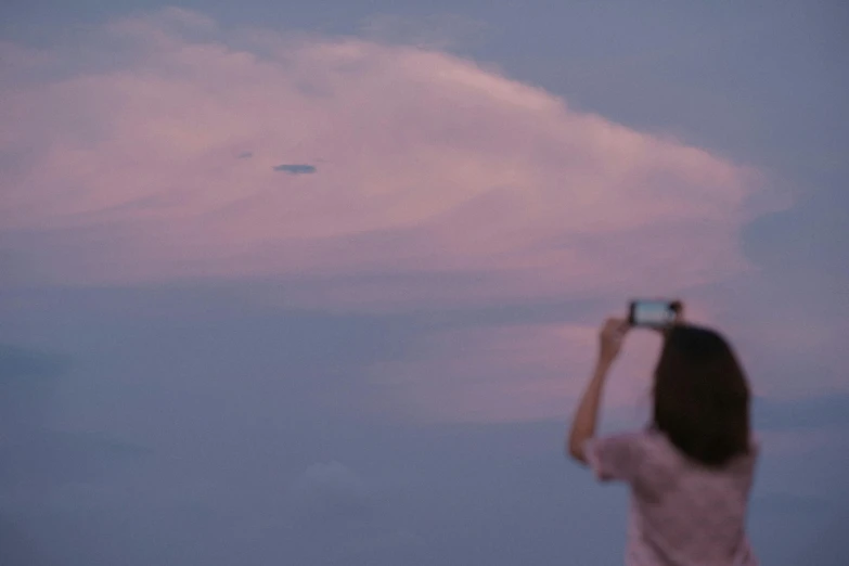 a woman taking a picture of a plane in the sky, inspired by Quint Buchholz, pexels contest winner, light pink clouds, flying saucer, 7 0 mm photo, at dusk!