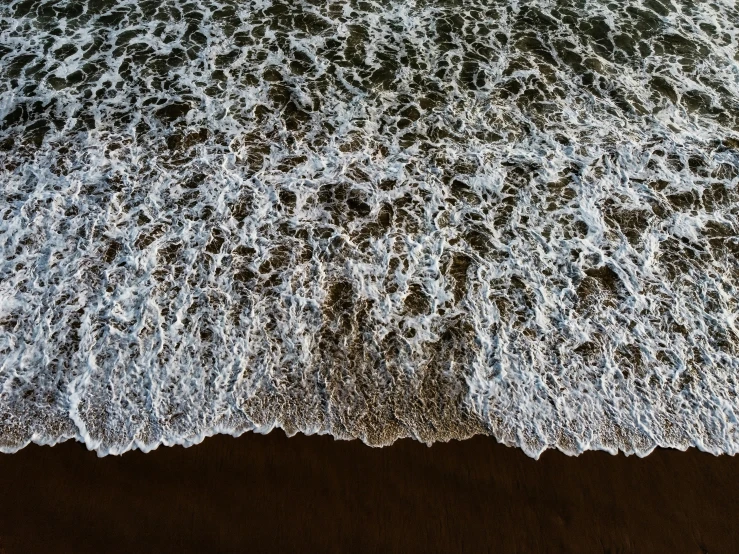 a person riding a surfboard on top of a sandy beach, inspired by Andreas Gursky, pexels contest winner, hurufiyya, extremely detailed water texture, view from the sky, black sand, rushing water
