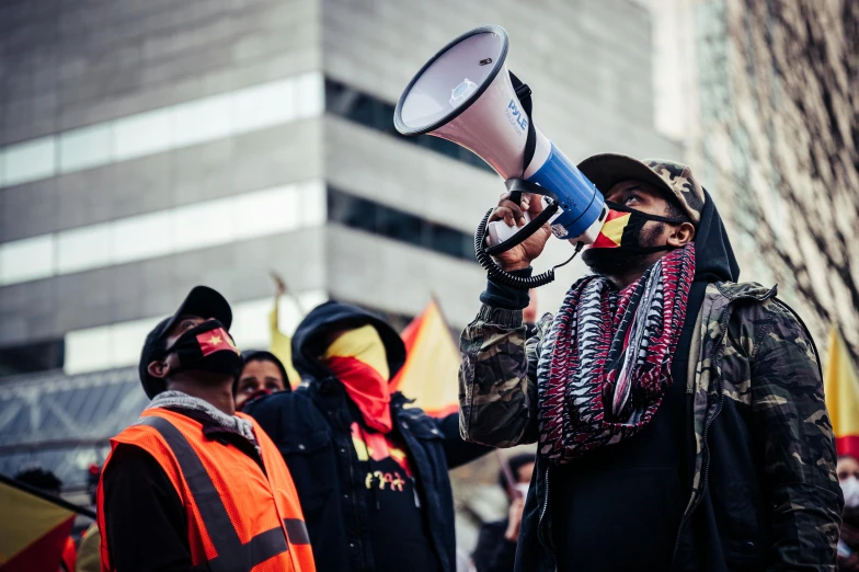 a man holding a megaphone in front of a crowd, a photo, by Julia Pishtar, pexels, indigenous man, black and yellow and red scheme, he wears dark visors, hoyte van hoytema