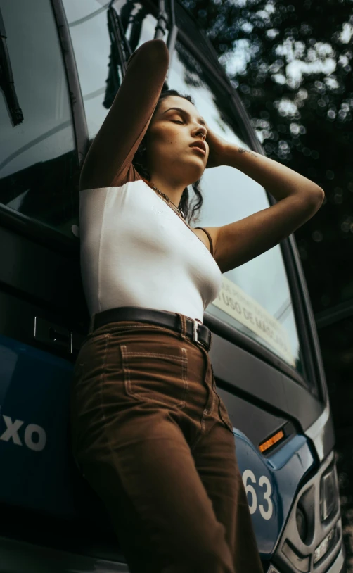 a woman leaning against the side of a bus, by Niko Henrichon, trending on pexels, renaissance, tight shirt, brown pants, official store photo, halter-top