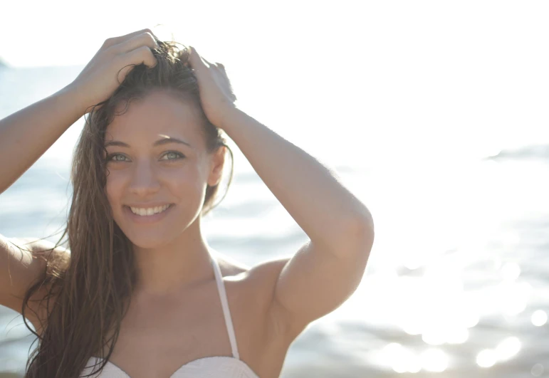 a woman standing on top of a beach next to the ocean, glowing skin face, smiling girl, hands in her hair, girl with brown hair