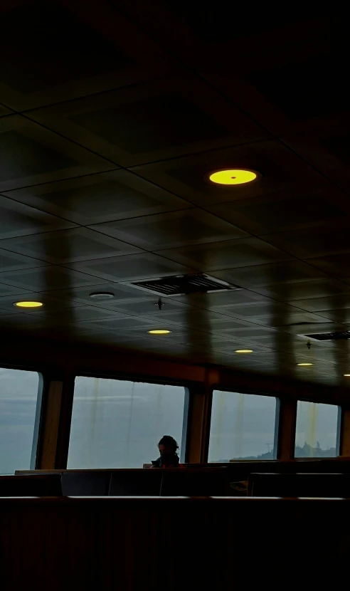 a man sitting at a table in front of a window, by Carey Morris, unsplash, interior of staten island ferry, ceiling hides in the dark, yellow lights, photo taken from a boat