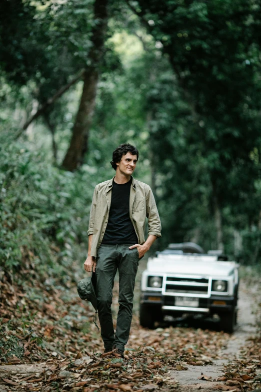 a man walking down a dirt road next to a car, an album cover, sumatraism, riccardo scamarcio, on a jungle forest, australian, profile picture